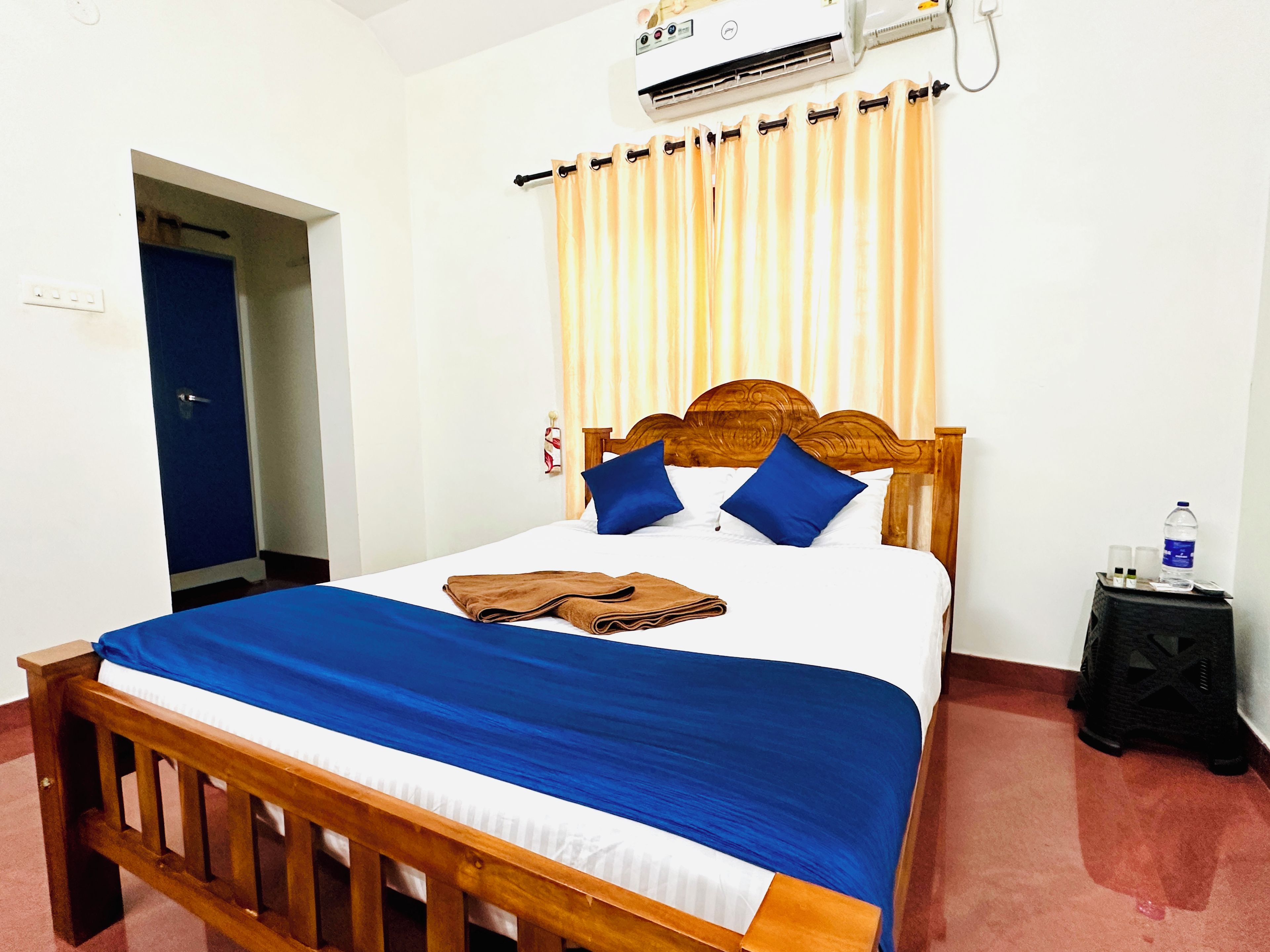 Habitación de hotel con cama de madera, colcha azul y blanca, cabecero de madera y unidad de aire acondicionado sobre la cama.
