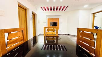 Pasillo de hotel con bancos de madera a ambos lados, una zona de estar central y un televisor montado en la pared.