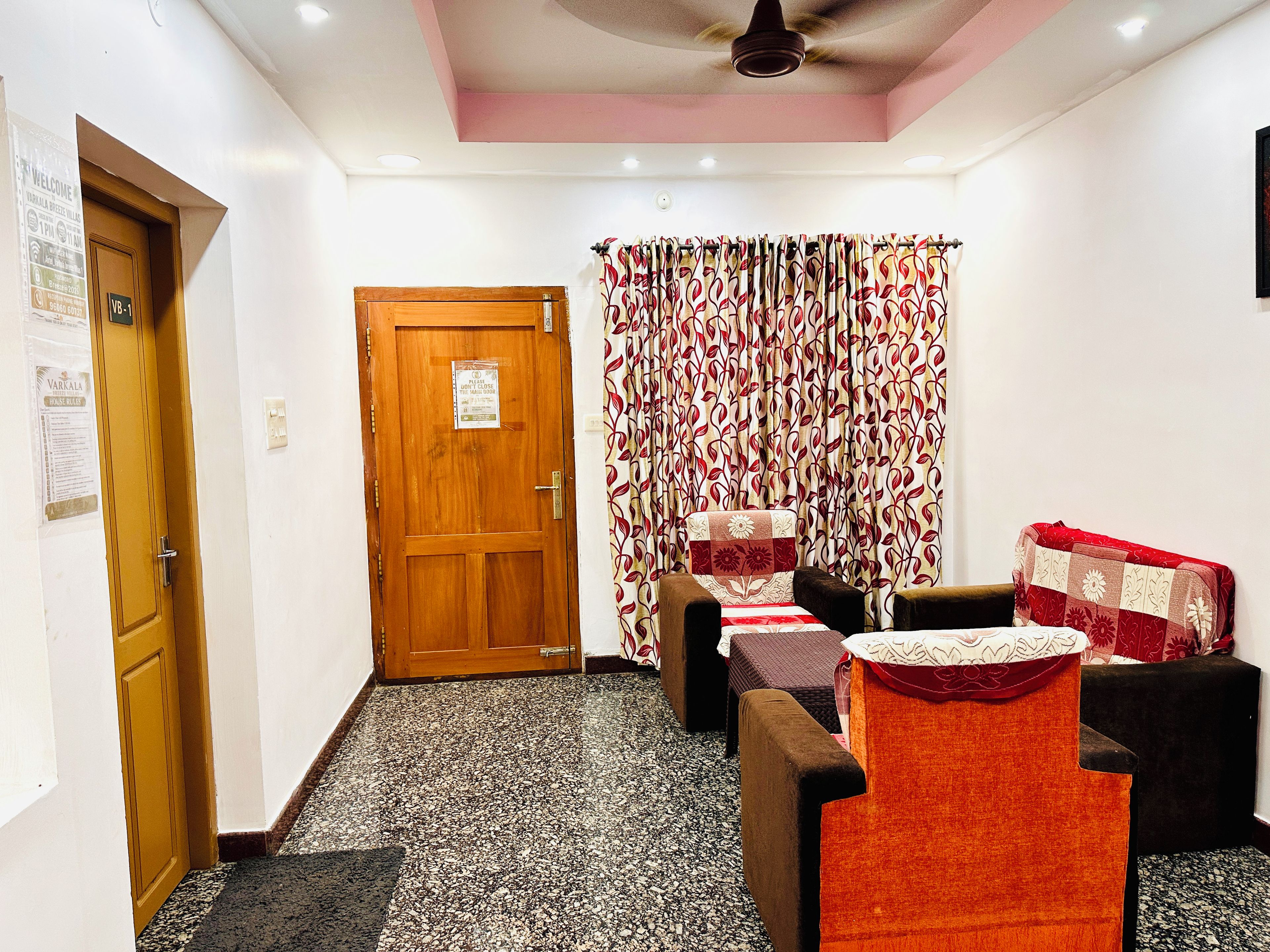 A room with a wooden door, a patterned curtain, a bed with a floral bedspread, and a small table with a red cushion.