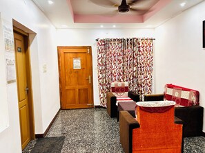 A room with a wooden door, a patterned curtain, a bed with a floral bedspread, and a small table with a red cushion. - Varkala Breeze Villas (Varkala)