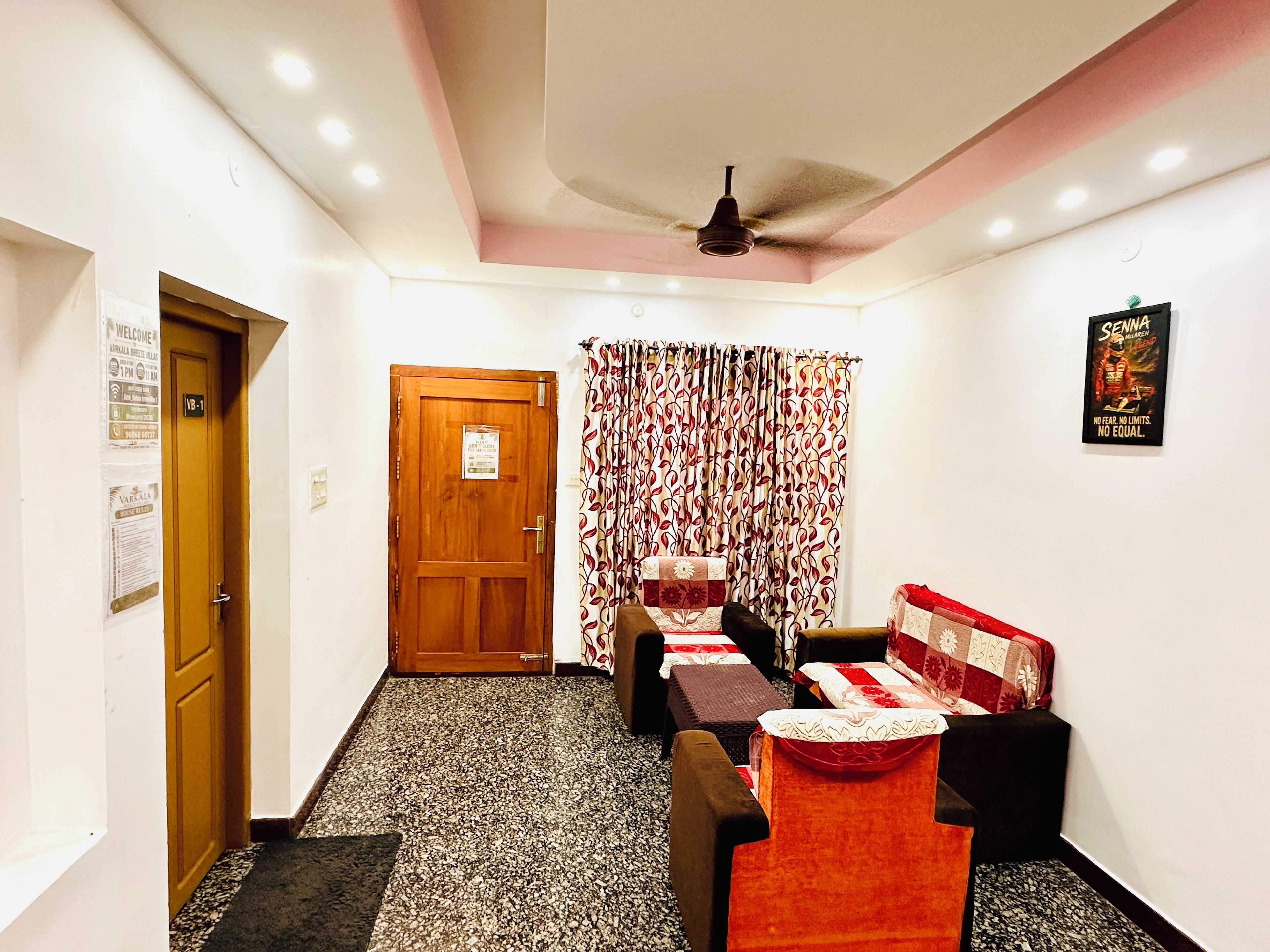 A hotel room with a wooden door, a ceiling fan, a patterned curtain, and a bed with a red and white quilt.