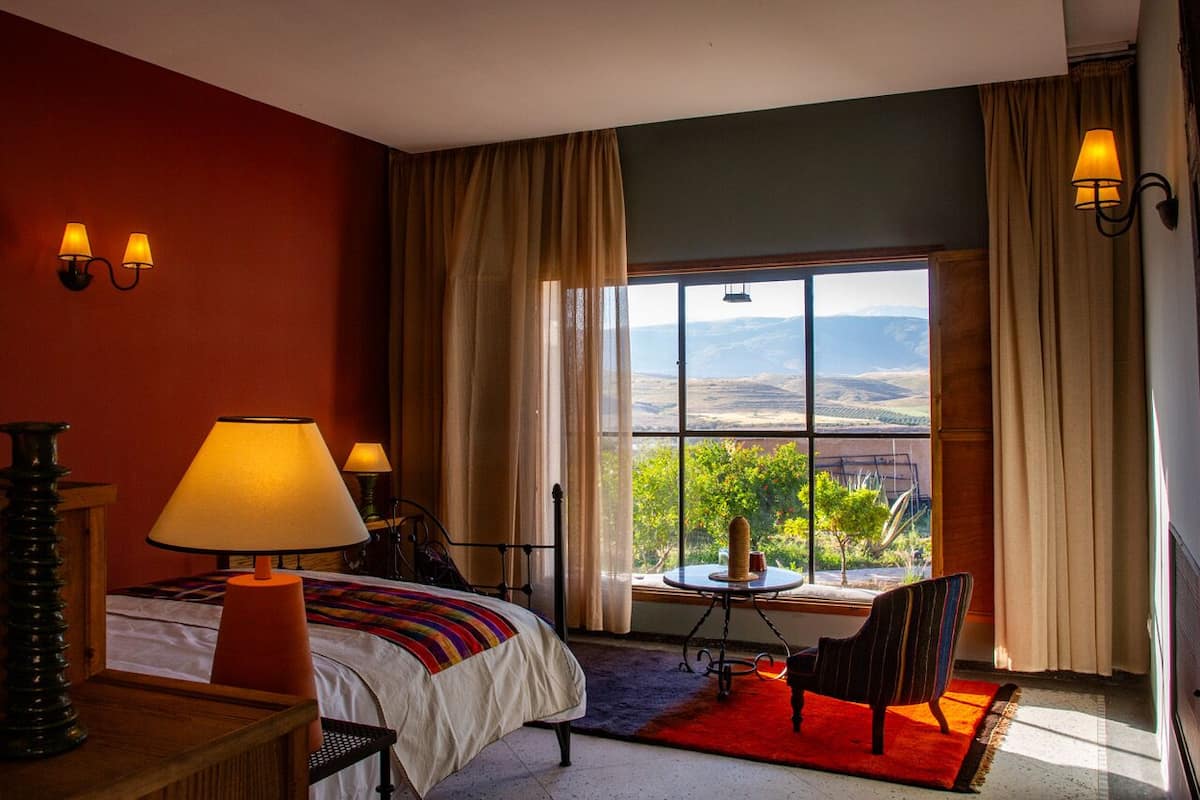 A hotel room with a bed, a table with a lamp, a chair, and a view of a mountainous landscape through large windows.