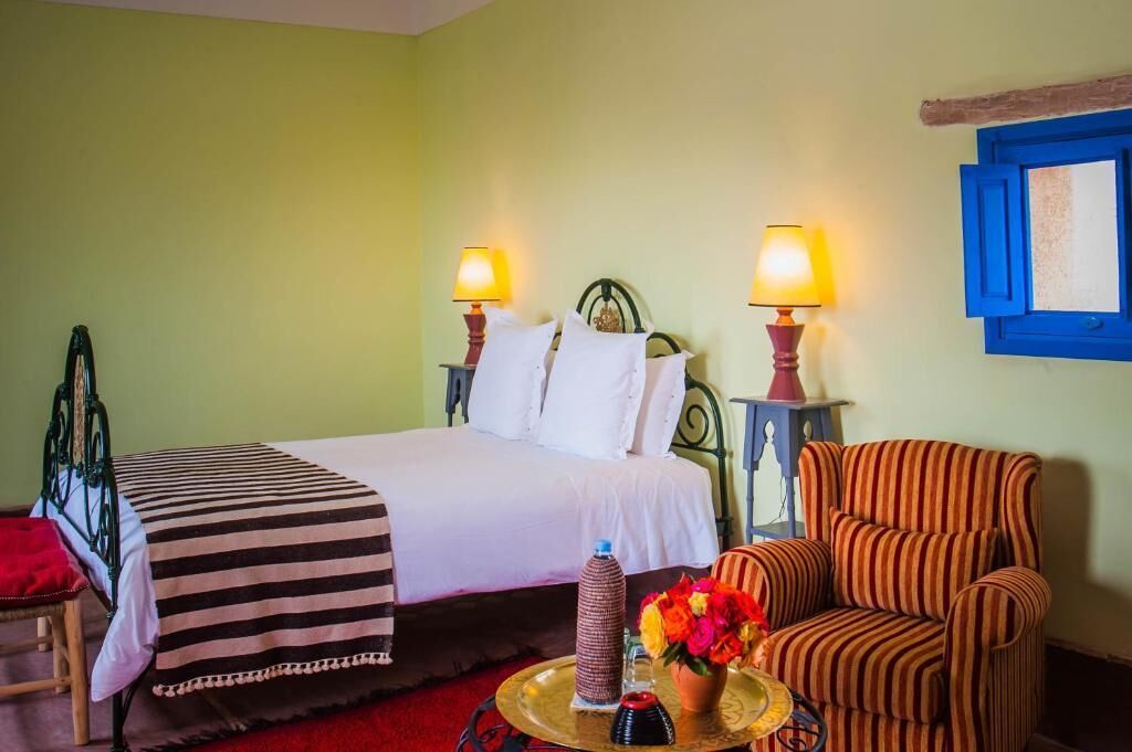 Una cama con sábanas blancas, dos lámparas de noche, un sillón a rayas y una mesita con un jarrón de flores.