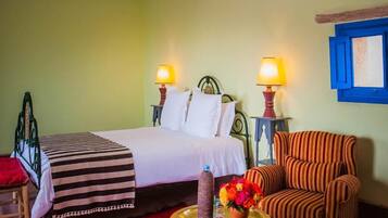 Una cama con sábanas blancas, dos lámparas de noche, un sillón a rayas y una mesita con un jarrón de flores.