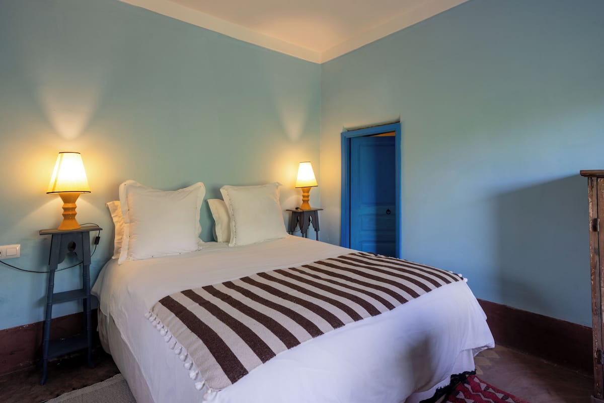 A bedroom with a bed, two bedside lamps, a blue door, and a small wooden nightstand.