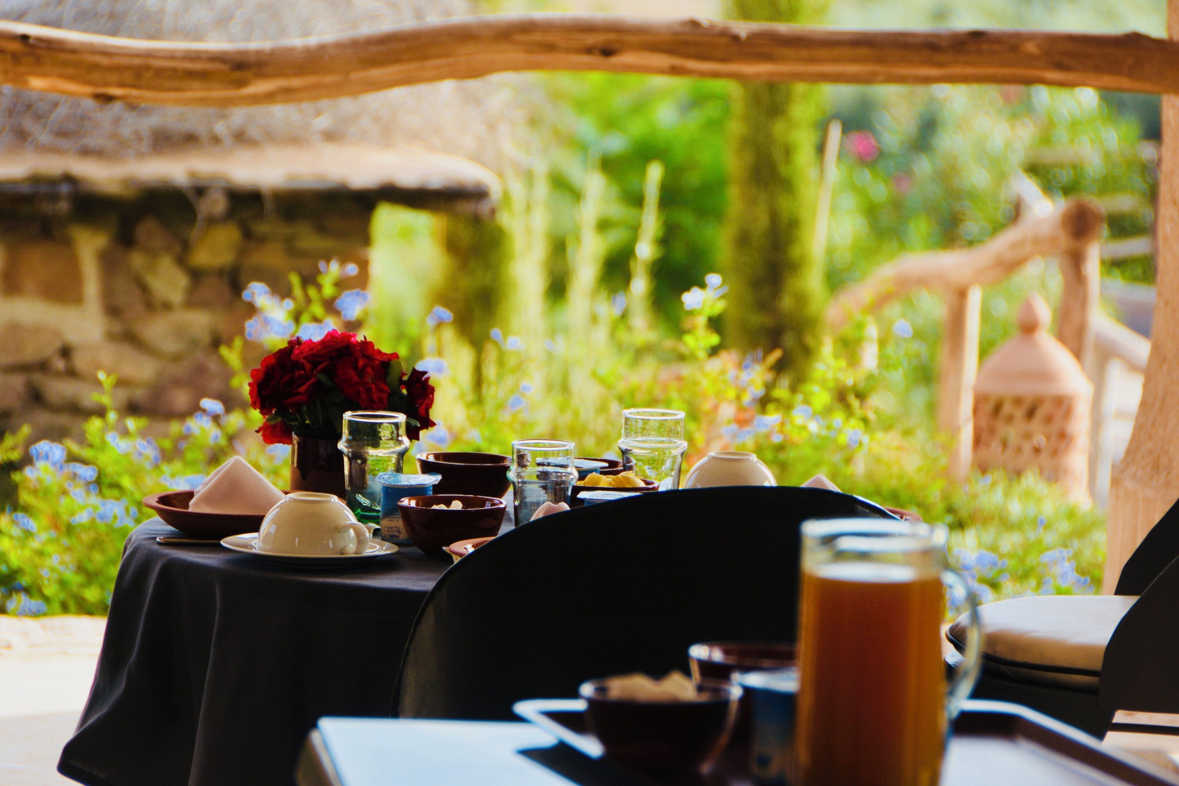 An outdoor dining area with a table set for two, surrounded by greenery and a stone wall.