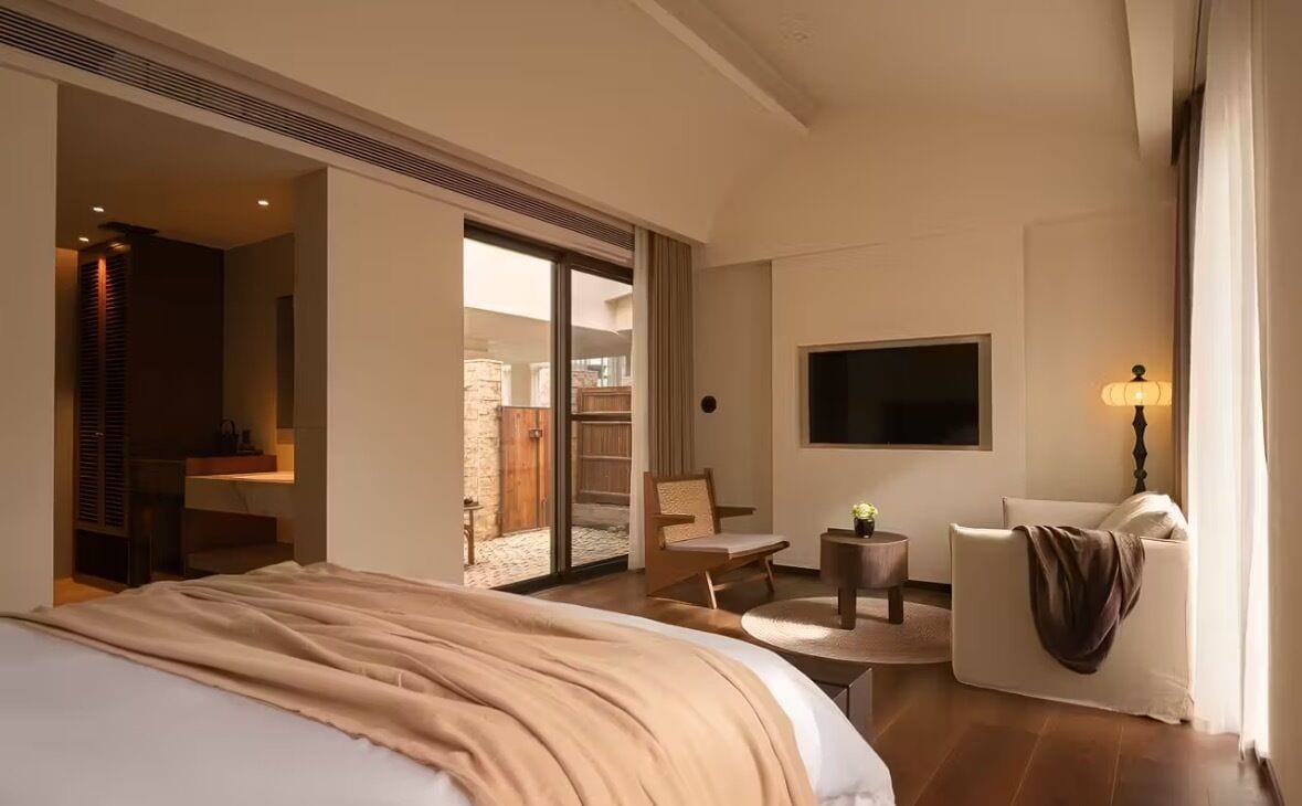 Habitación de hotel con cama, televisor, zona de estar con una silla y una mesa, y puerta corrediza de vidrio que da a un espacio exterior.