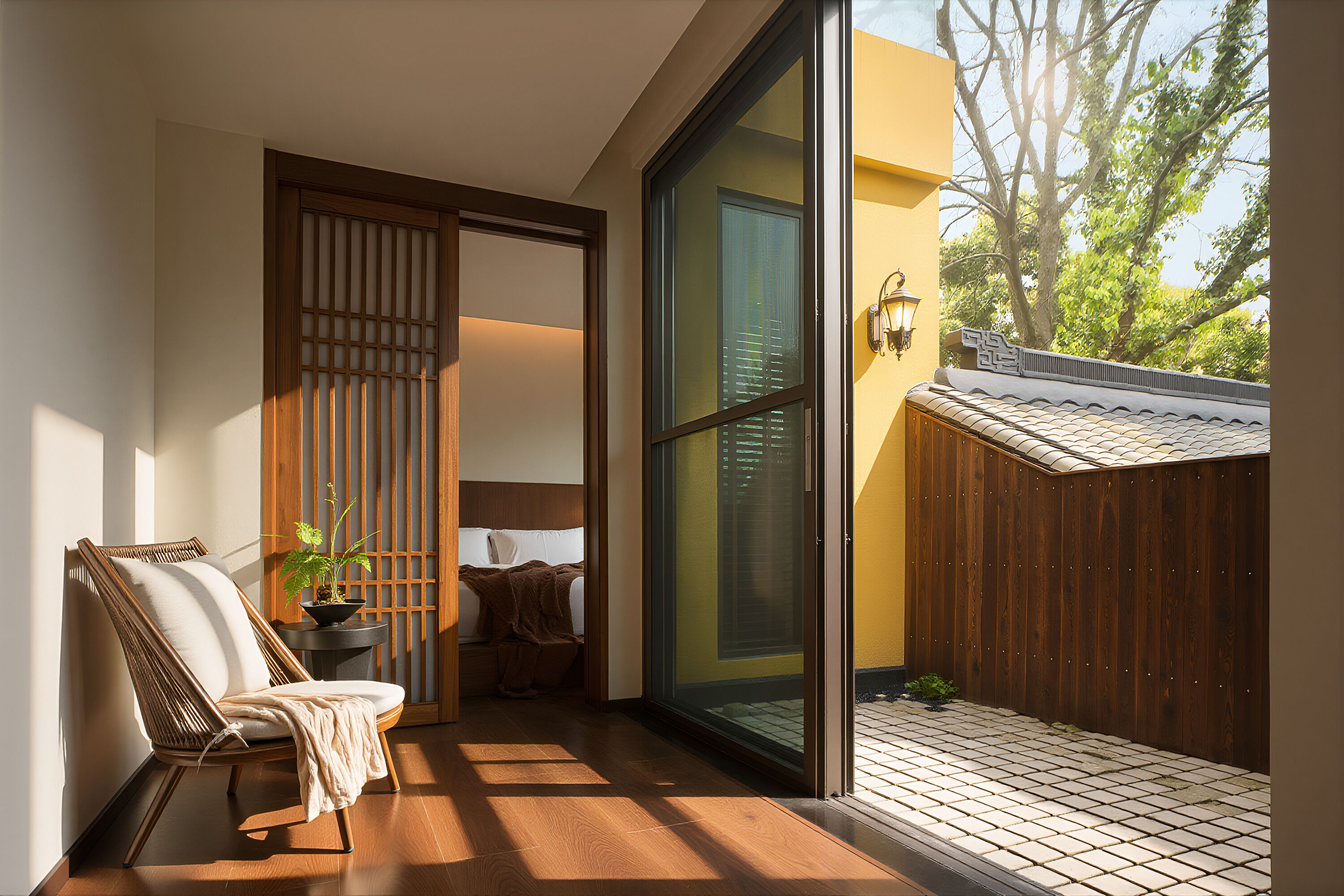 A room with a sliding glass door, a wicker chair, a small table with a plant, and a bed with a blanket.