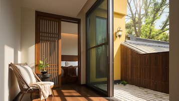 A room with a sliding glass door, a wicker chair, a small table with a plant, and a bed with a blanket.