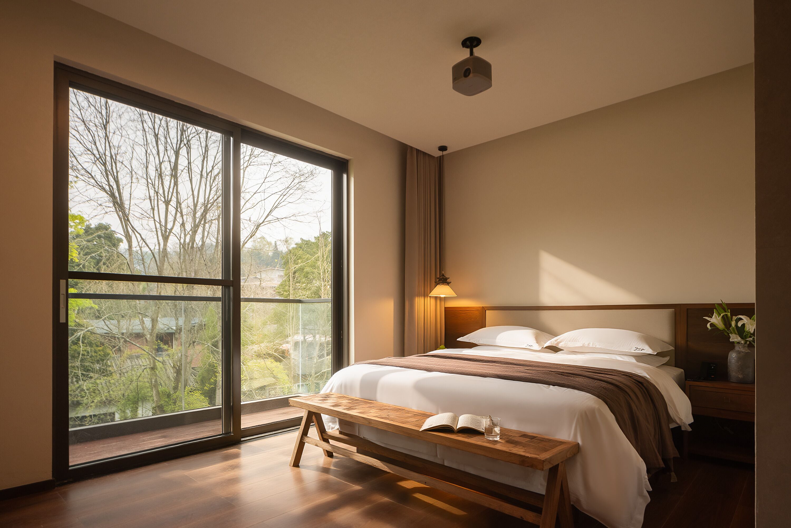 Ein Schlafzimmer mit großem Fenster, Nachttisch, Bank und Bett mit Kissen und Decke.