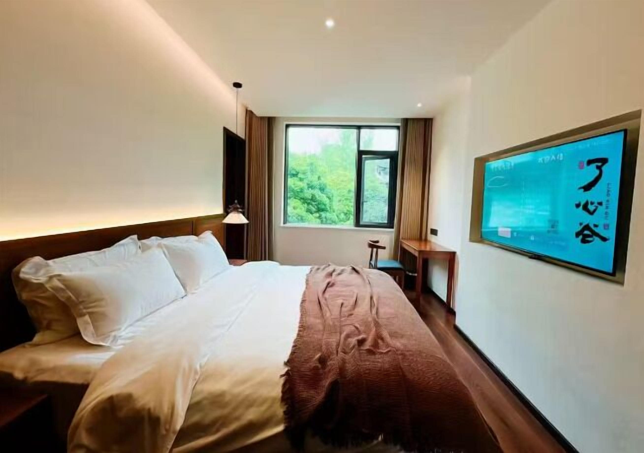 Ein Hotelzimmer mit Bett, einem Fenster mit Blick auf Grünflächen, einem an der Wand befestigten Fernseher und einem kleinen Tisch.
