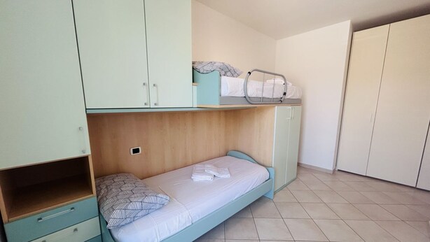 A single bed with a wooden headboard, a small cabinet with drawers, and a wardrobe with a mirror. - Viktor Flexrent - Abyssinia Real Estate (Riccione)