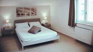 Ein Schlafzimmer mit einem Bett, zwei Nachttischen mit Lampen, einem Fenster mit Vorhängen und Wandbildern.