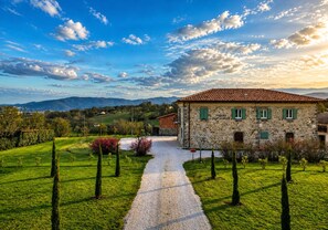 A stone building with a tiled roof, a gravel path, and a green landscape with trees.