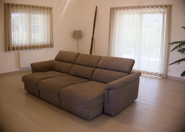 A living room with a brown sofa, two windows with sheer curtains, a floor lamp, and a radiator.