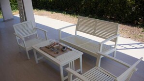 Une terrasse avec un banc blanc, deux chaises blanches et une petite table sur laquelle se trouvent un récipient en verre et des objets décoratifs.