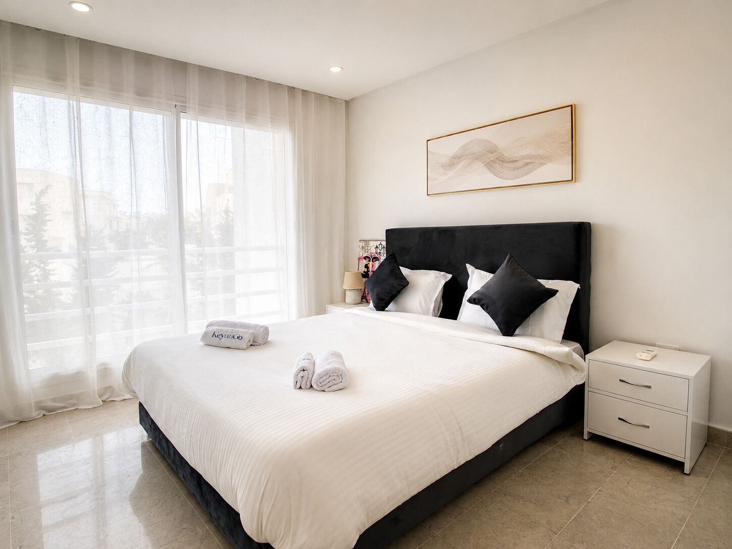 Una habitación de hotel con una cama grande, una mesita de noche, cortinas traslúcidas en la ventana y una obra de arte enmarcada en la pared.