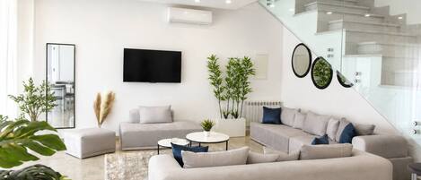 A modern living room with a large sectional sofa, a flat-screen TV, and a staircase with glass railings.