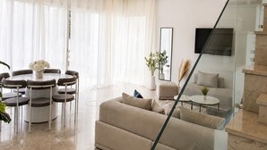 A modern living room with a beige sofa, a dining area with a round table and chairs, and a staircase with glass railings. - Elegant Villa in Gammarth With Private Indoor Pool (La Marsa)