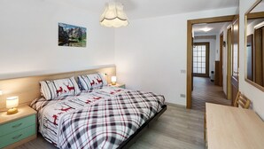 A bedroom with a bed, bedside tables, a hanging light fixture, a painting on the wall, and a hallway leading to another room. - Apartment 'Lella Genziana' with Mountain View, Balcony and Wi-Fi (Vigo di Fassa)