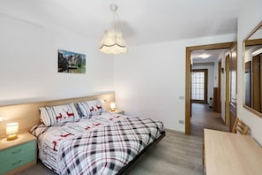 A bedroom with a bed, bedside tables, a hanging light fixture, a painting on the wall, and a hallway leading to another room. - Apartment 'Lella Genziana' with Mountain View, Balcony and Wi-Fi (Vigo di Fassa)