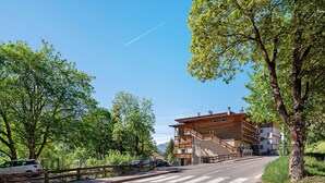 A road leading to a multi-story building with balconies, surrounded by trees and a clear sky. - Apartment 'Lella Genziana' with Mountain View, Balcony and Wi-Fi (Vigo di Fassa)