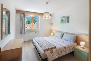 Ein Schlafzimmer mit Bett, Nachttischen, Schreibtisch, Fenster mit Vorhängen, einer Pendelleuchte und einem Wandbild.