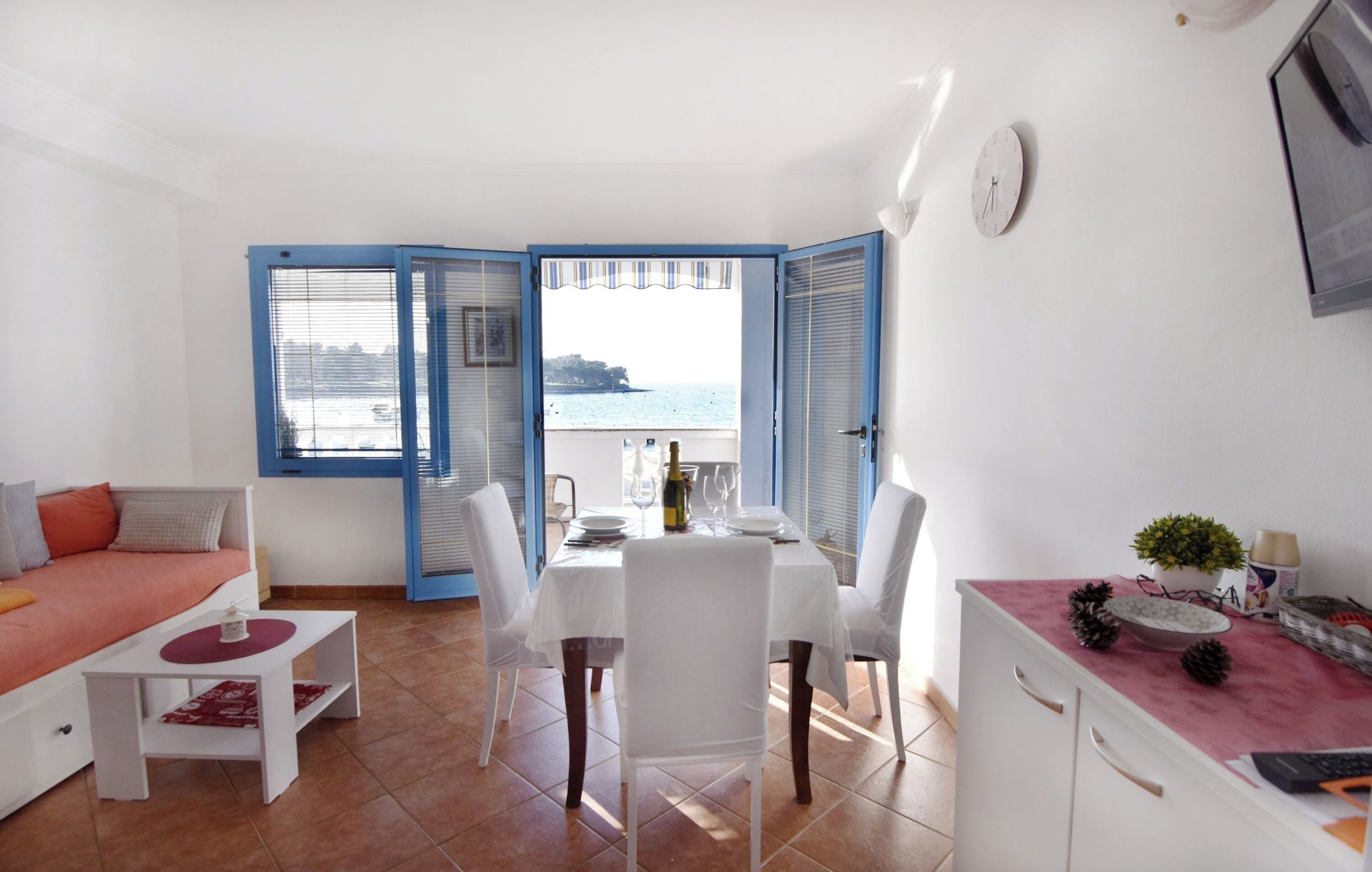 A room with a dining table, chairs, a sofa, a small table with a red placemat, a clock on the wall, and a view of the sea through blue shutters.