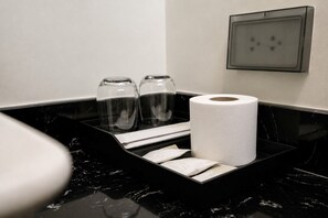 Une salle de bain avec un comptoir en marbre noir, des toilettes, deux bouteilles en verre transparent, un rouleau de papier toilette et un distributeur de serviettes en papier.