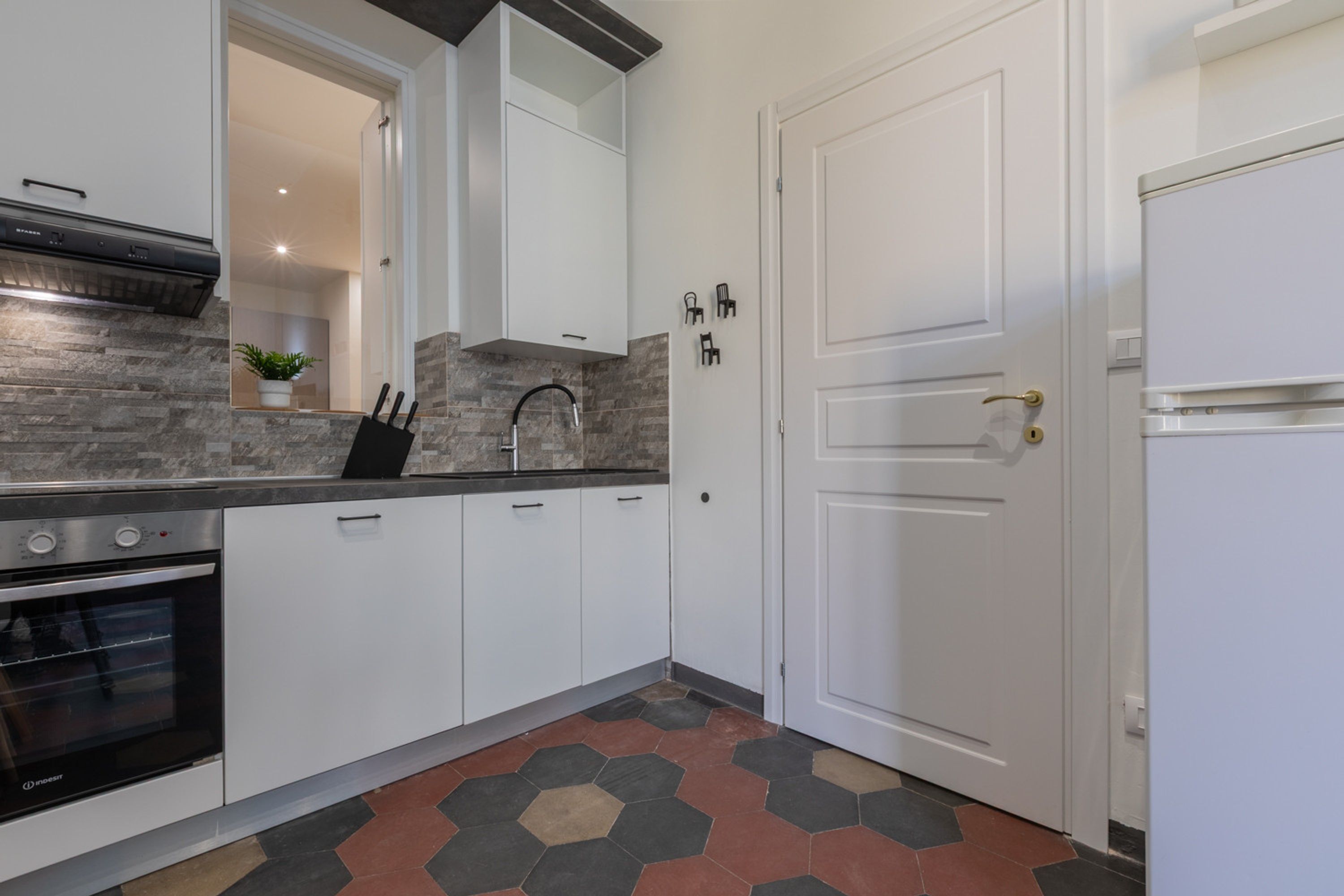 A modern kitchen with white cabinets, a built-in oven, a stainless steel range hood, a hexagonal patterned floor, and a white door with a gold handle.