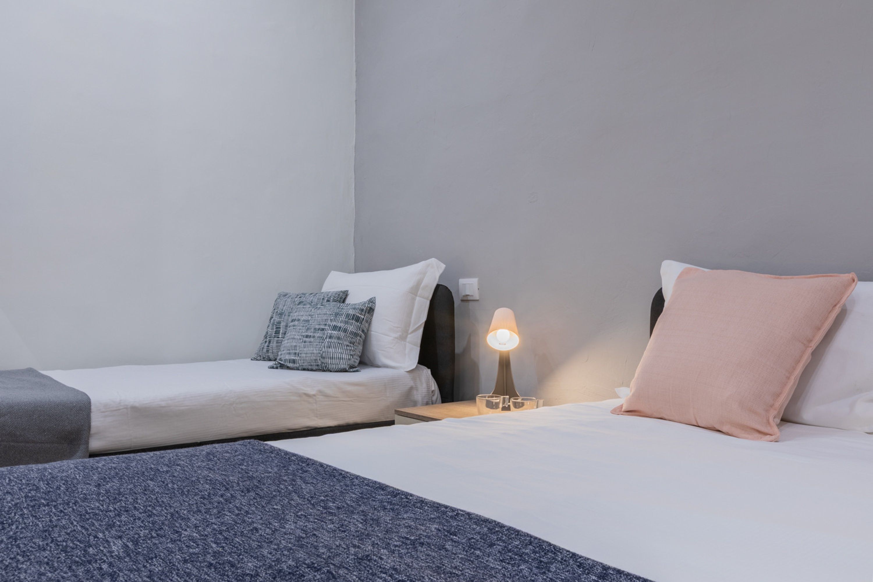 A hotel room with two single beds, a nightstand with a lamp, and a small table with glasses.