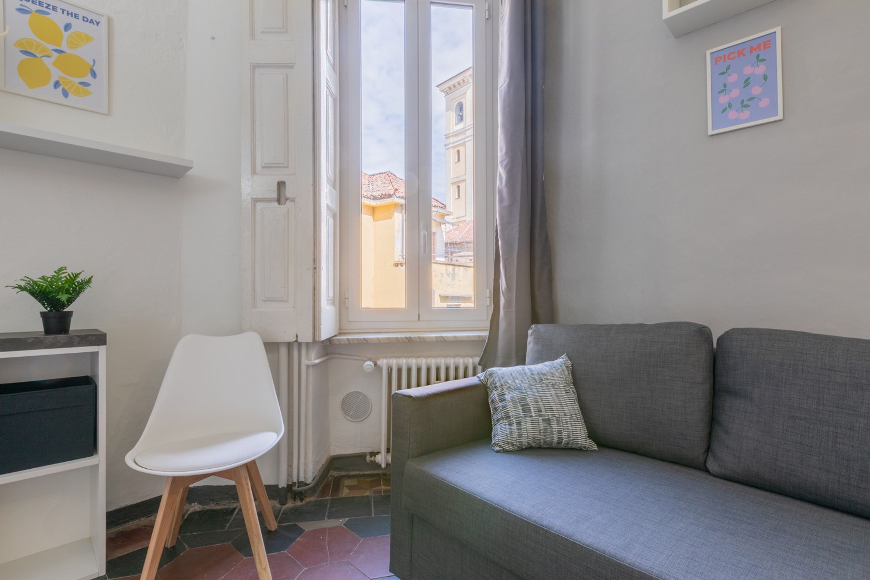 Una stanza con un divano grigio, una sedia bianca, un termosifone, una finestra che si affaccia su edifici e una pianta su uno scaffale.