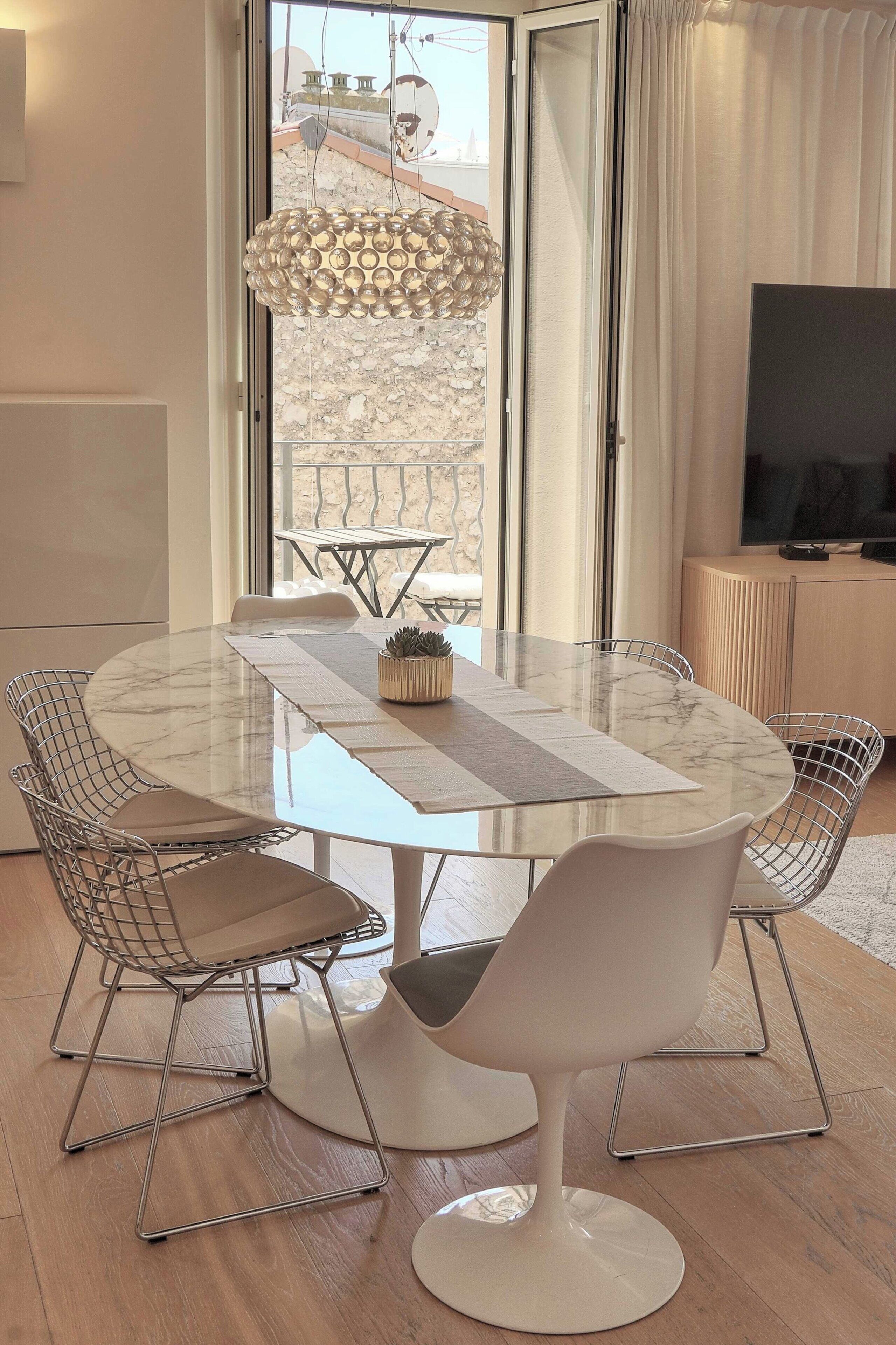 A round dining table with wireframe chairs, a marble top, and a hanging light fixture.
