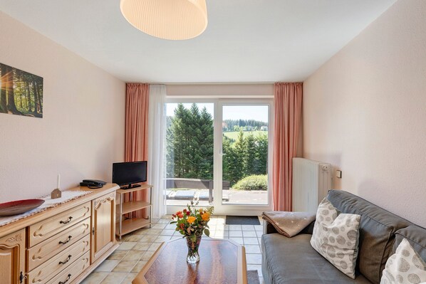 Ein Wohnzimmer mit einer Couch, einem Couchtisch, einem Fernseher, einem Schrank und einem großen Fenster mit Vorhängen.