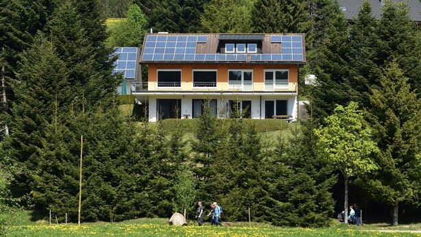 A two-story house with solar panels on the roof, surrounded by dense forest. - Apartment Tanne – Opposite Ski Lift with Private Terrace, Garden, and Green Views (Schönwald im Schwarzwald)