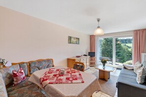 Ein gemütliches Zimmer mit einer Couch, einem Tisch mit einer Stegdecke, einem Schreibtisch mit einem Computer und Blick auf Grünfläche draußen.
