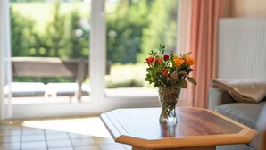 A room with a wooden table, a vase with flowers, a sofa, and a view of greenery outside. - Apartment Tanne – Opposite Ski Lift with Private Terrace, Garden, and Green Views (Schönwald im Schwarzwald)