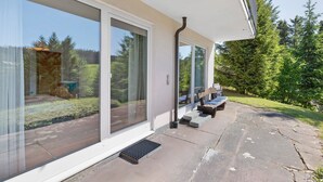 A modern house with large sliding glass doors, a stone patio, and a small outdoor seating area. - Apartment Tanne – Opposite Ski Lift with Private Terrace, Garden, and Green Views (Schönwald im Schwarzwald)