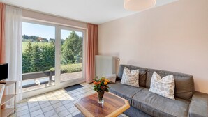 A living room with a sofa, a coffee table, a TV, and a view of a green landscape through large windows. - Apartment Tanne – Opposite Ski Lift with Private Terrace, Garden, and Green Views (Schönwald im Schwarzwald)