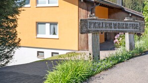 Un letrero de madera con el texto 'FERTENWOBENBURG' grabado, un edificio amarillo con techo marrón y un paisaje verde.