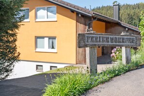 Et træskilt med 'FERTENWOBENBURG' udskåret, en gul bygning med et brunt tag og et grønt landskab.