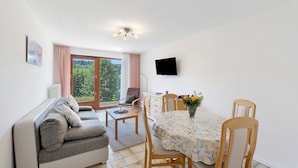 Sala de estar con sofá, mesa de comedor, sillas, televisor y vista a zonas verdes por la ventana.