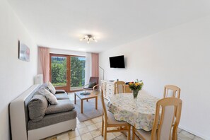 En stue med sofa, spisebord, stole, fjernsyn og grønne omgivelser udenfor vinduet.