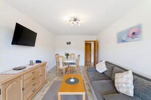 A living room with a flat-screen TV, a wooden dining table, a sofa, and a painting on the wall. - Charming Apartment Birke – Opposite Ski Lift with Private Terrace, Garden & Green Views (Schönwald im Schwarzwald)