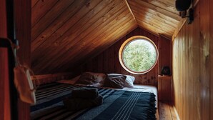 A cozy attic bedroom with a sloped ceiling, a round window, a bed with striped bedding, and wooden walls.