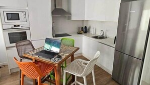 Een kleine keuken met witte kasten, een roestvrijstalen koelkast, een magnetron en een laptop op een tafel met kleurrijke stoelen.