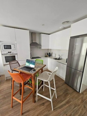 Eine kompakte Küche mit weißen Schränken, einem Edelstahlkühlschrank, einer Mikrowelle und einem Laptop auf einem Tisch mit bunten Stühlen.
