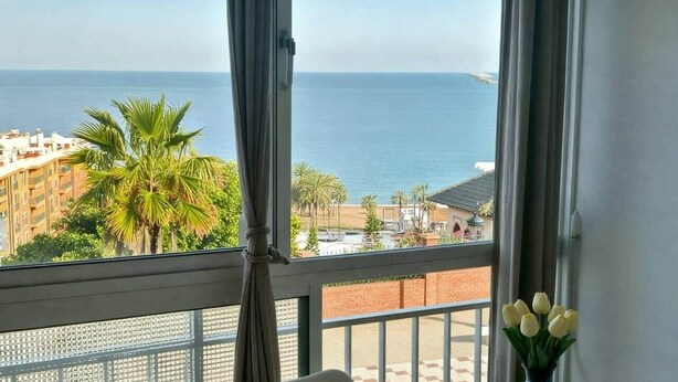Uma varanda com uma mesa, uma tigela de fruta, um vaso com flores e vista para o mar e edifícios.