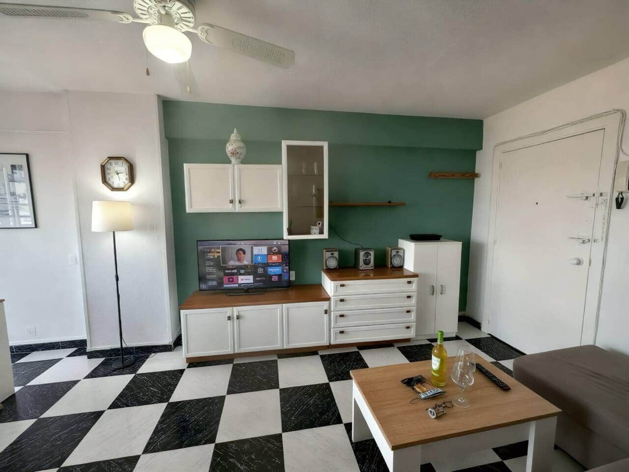 Een kamer met een geruite vloer, een houten tafel met een fles en glazen, een lamp, een televisie waarop iemand te zien is, en een witte kast met lades.