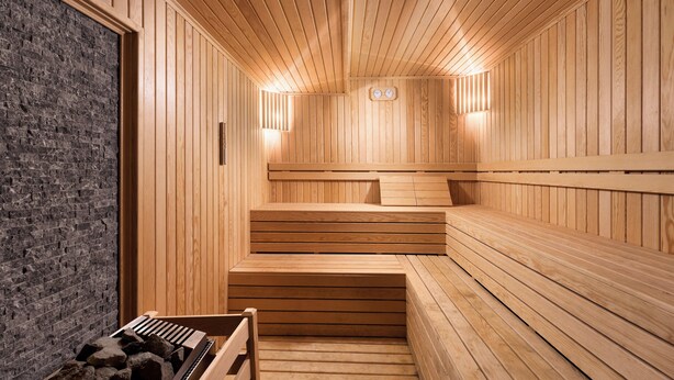 Un sauna avec des murs et des bancs en bois, un mur de pierre d’un côté et deux lampes fixées au mur.
